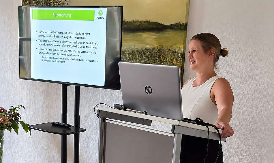 Dr. Neele Riess hält einen Vortrag und steht vor einem Bildschirm mit Powerpoint-Präsentation. 